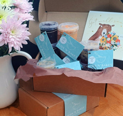 Box of six puddings in little pots with blue labels,  on top of another box of the same size which has a blue band wrapped around it. Jug of pink and yellow flowers on left hand side.  There is a card with Get Well on it and a bear holding a bunch of flowers.